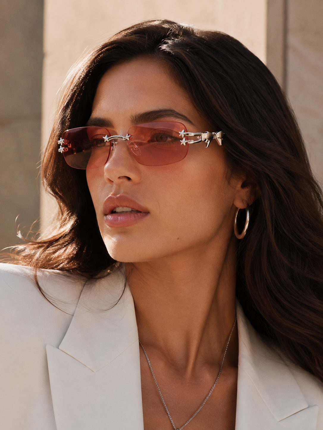 A close-up of a woman wearing rimless sunglasses with a pink-tinted lens and silver decorative metal frames, styled with a white blazer and gold hoop earrings.